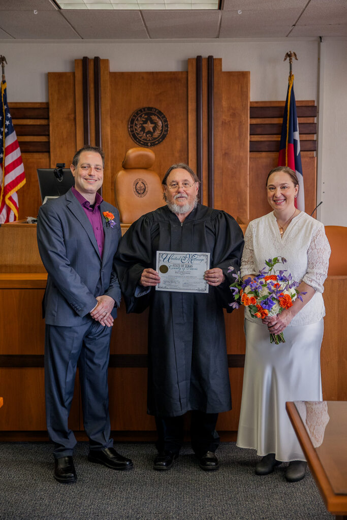 Wes and Rachele stand with the judge in the courtroom as he holds up the signed marriage license.