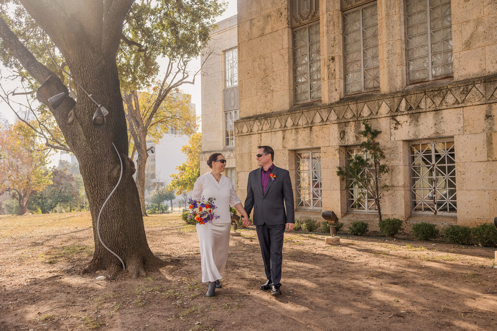 Outside the courthouse, Rachele and Wes walk hand in hand in the shade of a large oak tree.
