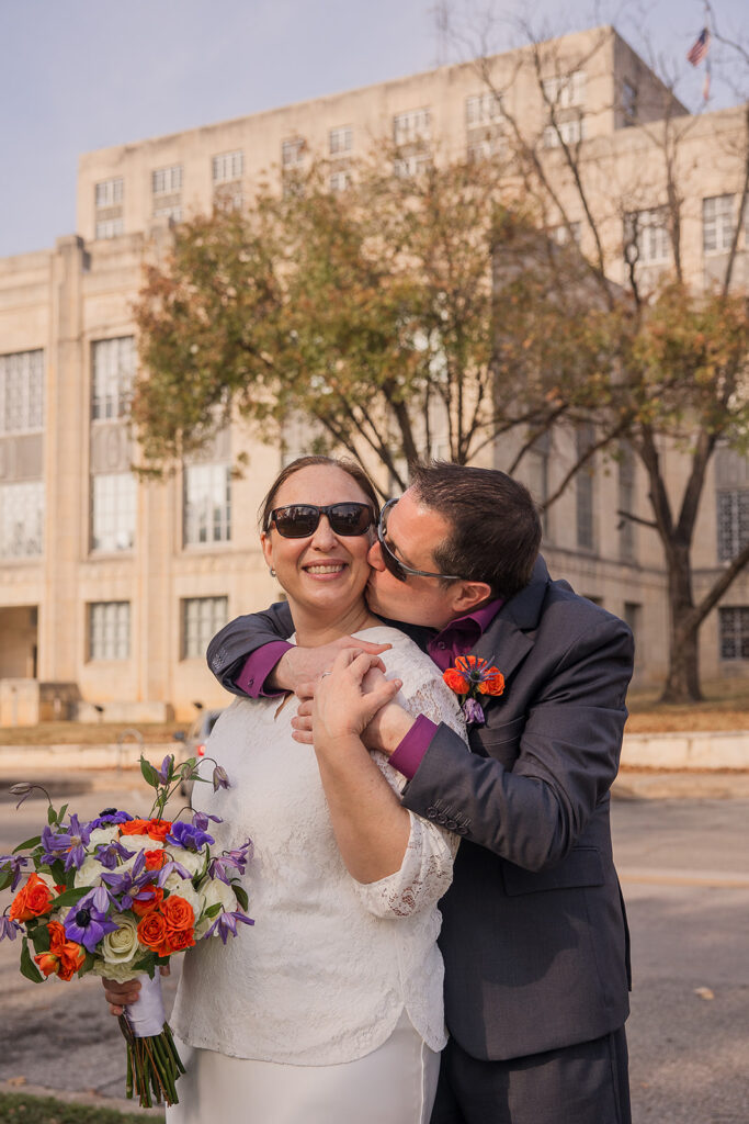 Wes kisses Rachele on the cheek from behind with the facade of the Travis County Courthouse behind them.