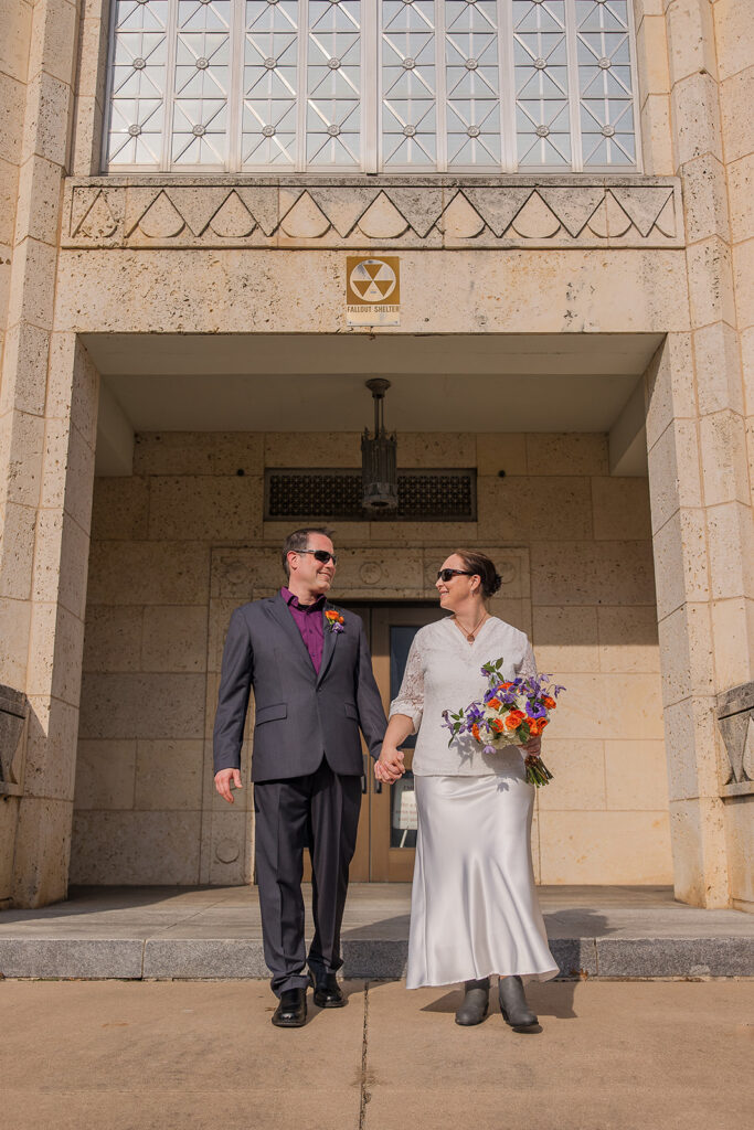 Wes and Rachele stand hand in hand outside the Travis County Courthouse side entrance with an old fallout shelter sign above them.