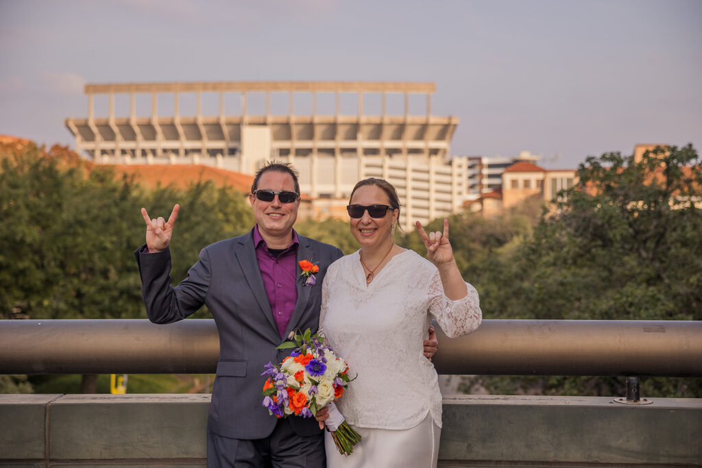 Wes and Rachele hold up their hook 'em horns hand signals as they stand together with DKR memorial stadium behind them.