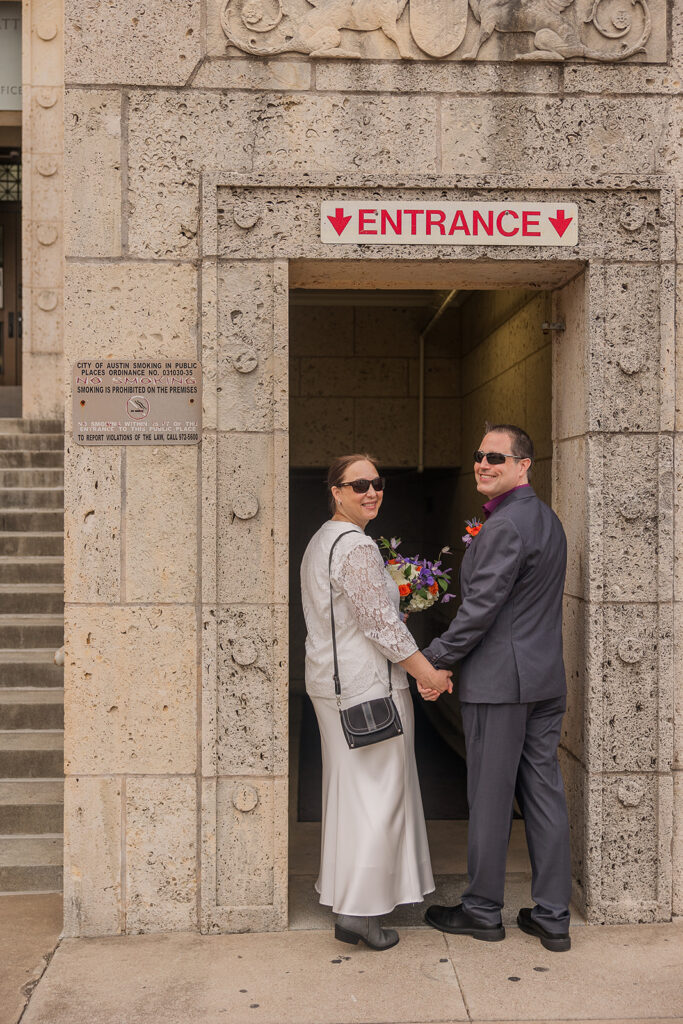 Rachele and Wes hold hands as they pass through the entrance way into the courthouse. They look back over their shoulders, smiling.