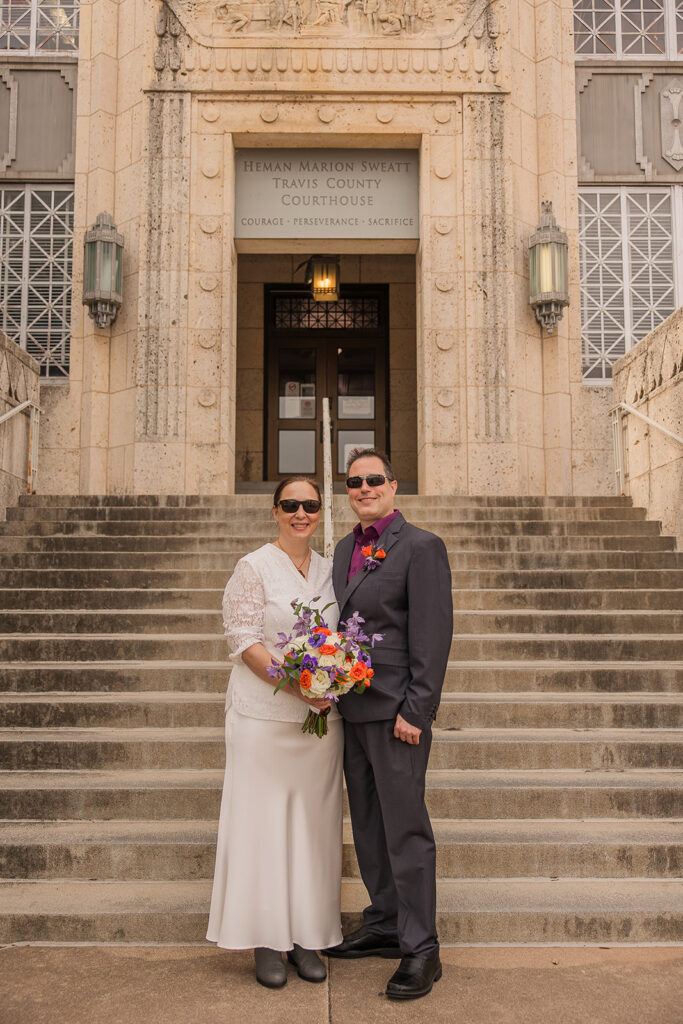 Rachele and Wes on their wedding day standing in front of the steps up to the Travis County Courthouse, a limestone-clad building in the PWA Moderne style of the 1930s. Rachele is wearing a white lace blouse and long white satin skirt with gray boots. Wes is wearing a dark gray suit and eggplant purple shirt. Rachele is holding her bouquet of white and orange roses with purple flowers mixed in. Both of them are wearing their sunglasses.