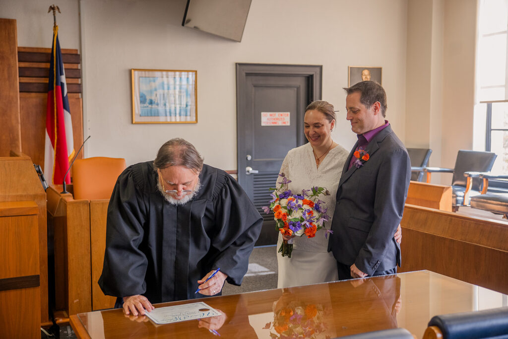 Rachele and Wes watch as the judge signs their marriage license in the court room.