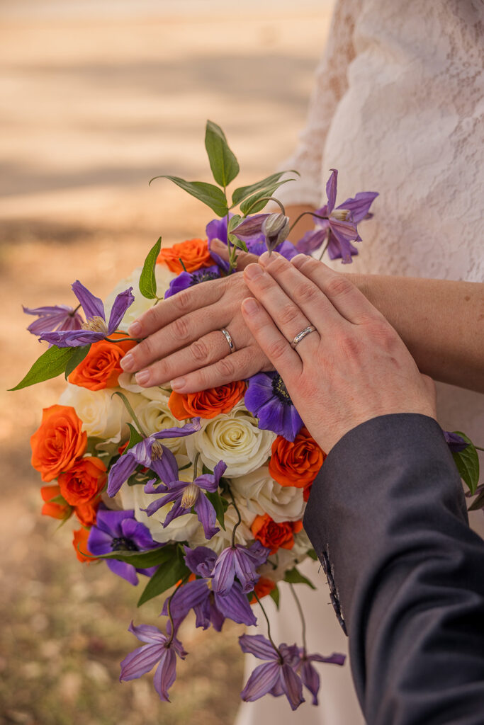 A close up of Rachele and Wes’s left hands showing their matching plain white gold wedding bands with Rachele’s bouquet of white and orange roses with purple flower highlights behind their hands